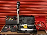 Overview of the lot showing multiple items grouping - laser level kit with tripod and yellow tape, hand tool set in black case, red case with jumper cables.