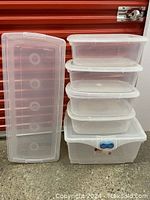 Five clear plastic storage tubs, one large with blue latch and four smaller identical tubs stacked, placed in front of red roll-up door.