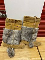 Photo shows a pair of tan mukluk boots with wide bands of gray faux fur on the front, decorative beaded pattern near the top, and tan suede exterior with stitching details and pom-pom laces.