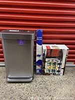 Lot showing the Simplehuman stainless steel waste can with visible dents and scrapes on the lid and back, alongside the boxed new stackable dryer rack and can storage rack.