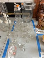 Multiple clear glass tumblers and shot glasses arranged on table alongside large glass ice bucket and tall cocktail shaker with metal lid.