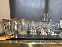 Overview of 16 drink glasses: 5 short water glasses with gold rim on left, 7 stemmed brandy glasses in center-right, 2 smaller sherry glasses also stemmed near center.