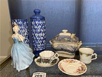 Lot showing two tall blue and white ceramic canisters (one with lid), ceramic figurine in blue dress, blue and white sugar bowl, floral teacup with matching saucer, teacup with chip, and an extra saucer.