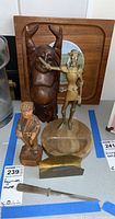Four figurines on a table: a carved face, wooden man with fishing net, brass man playing horn, carved wooden pipe smoker.