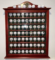 Wooden and glass door golf ball display case with 56 golf balls arranged inside on shelves, featuring various golf-related logos.