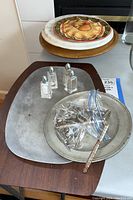 Tin tray with three glass salt and pepper shakers, metal tray underneath, small metal dish holding bag with metal corn holders and handle, stacked plates behind on table.