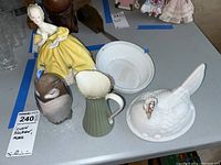 Porcelain and ceramic figurines and Lenox pitcher on table with blue tape notes for chips
