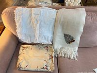 Three linen items including white pillow shams, a wool shawl with embroidered Christmas tree, and a folded Christmas-themed table runner showing holly and berries.