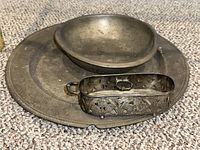 Wide shot showing all three metal items: round platter, oval bowl, and decorative holder with broken handle placed on carpeted surface.