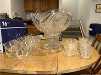 Shot of punch bowl on matching pressed glass stand with rows of 19 glass tumblers on left and 8 pressed glass cups on right on wooden table