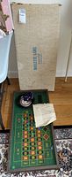 Full view of cardboard box labeled 'roulette game' beside the roulette board with wheel and instruction sheet on a hardwood floor.