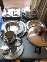 Overview of silverplate serving pieces including trays, covered serving dishes, copper chafing dish on stand, and silverplate pitchers.