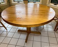 Round wooden table on a tiled floor, visible surface stains and wood grain, central pedestal base with four feet