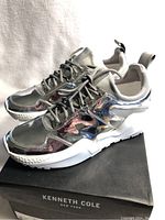 Side angled view of the sneakers on top of the Kenneth Cole branded black box, showing the silver metallic reflective finish and sole detail.