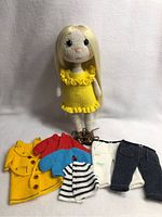 Handmade crochet doll dressed in a yellow ruffled dress standing behind various crochet and knit clothing items on a white background.