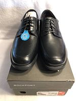 Front view of black men's Rockport dress shoes, size 9, on the box showing lace-up design and polished finish.
