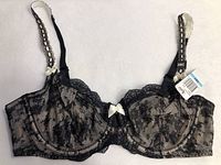 Photo showing the black lace bra with nude lining, white bows, and size tag 38C.
