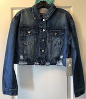 Front view of a medium blue women's cropped denim jacket showing distressing on front hem with buttons and chest pockets visible.