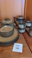 Full view of the Denby pottery set showing stacked plates, cups, a teapot, creamer, and casserole on a wooden surface.