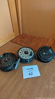 Three used fly fishing reels lined up on a wooden surface, with a MaxSold auction card labeled 42 in front.