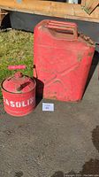 Two red metal gas cans placed on ground, one large rectangular and one smaller cylindrical marked 'GASOLINE'