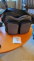 Closed black and gray camera bag with multiple zippered compartments and adjustable shoulder strap on a wooden table with auction tag.