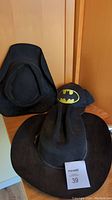 Three black hats displayed together, including a wide-brimmed western hat, a smaller tattooed western hat, and a Batman logo hat.