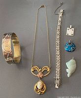 Full lot overview showing mixed metal bangle, gold colored Victorian style pendant with cameo, silver bracelet, rectangular silver pendant with bail, blue enamelled round pendant with colored stones, and green stone fish pendant.