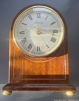 Front view of the Quincy mantel clock showing the round clock face with Roman numerals, gold hands, and wooden curved body resting on metal feet.
