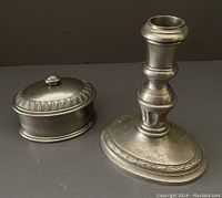 Pewter oval lidded box and candlestick shown side by side on flat surface