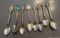Top view showing 8 sterling silver souvenir spoons with detailed enamelled crests of various colors and designs on the handle tops.