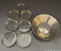 Seven round coasters with sterling silver rims and etched glass bases arranged in a scattered formation with one modern sterling silver bowl beside them.