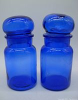 Front view of two cobalt blue glass apothecary jars with lids on, showing size and shape.