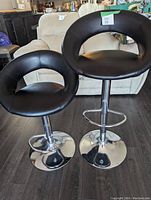 Two black faux leather swivel bar stools with silver metal bases shown from side angle on dark wood floor.