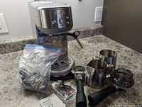 Photo of the Breville Bambino espresso maker with steam wand and accessories including milk pitchers, tamper, portafilter, and cleaning tablets on countertop.