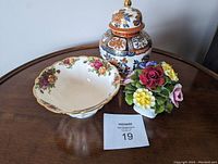 Three decorative china pieces on wooden table: ginger jar, floral pedestal bowl, floral figurine with auction tag 19