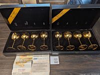 Two black presentation boxes each holding four gold-tone wine goblets with satin lining. A vintage magazine ad and informational paper are placed in front.