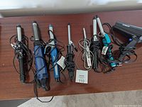 Photo showing six curling irons of various sizes and one black Conair hair dryer on a wooden surface.