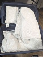Blue plastic tote filled with several white lace curtain panels showing intricate floral lace patterns, some folded neatly.