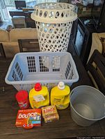 Front view showing tall laundry basket behind rectangular basket with detergents, spray, laundry bars, and bucket in front