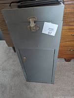 Gray metal locking security cabinet with a latch on top and a lockable lower door, positioned on carpeted floor near wooden furniture.