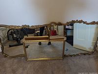 Three gold-framed mirrors on carpeted floor: two large ornate curved mirrors behind smaller rectangular mirror with gold beaded frame.