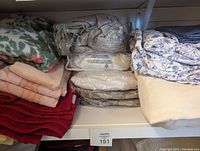 Shelf with assorted folded towels in multiple colors including red, yellow, pink, and floral patterns, along with packaged sheets and mattress pads wrapped in plastic.