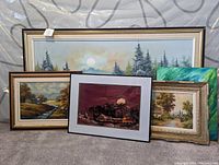 Five assorted wall art pieces including framed landscapes, one textile piece and a bird painting, displayed together on carpet against an interior backdrop.