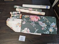 All wallpaper rolls spread on table showing variety including green floral with tear, white floral, silver roll, and green border trim rolls.