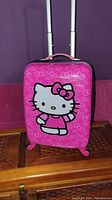 Front view of bright pink child-sized carry on with Hello Kitty large face graphic and bow pattern.