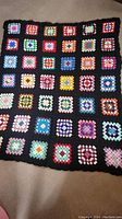 Handknitted multicolor square patch blanket throw laid out, showing rows of granny square colored patches with black borders.