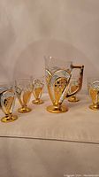 Full view of the vintage glass pitcher in the center with five matching glasses arranged around it. All have gold pineapple pattern and gold bases.