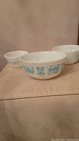 Three white Pyrex mixing bowls with blue Amish pattern visible on the outside. The bowls are arranged from smallest to largest.