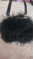 Front side view of a small black feather purse on a beige surface, showing its soft fluffy texture, black strap and gold-tone clasp.